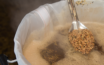 Close-up of a spoon lifting wet crushed grains from a mesh brew bag, showing wort beneath.