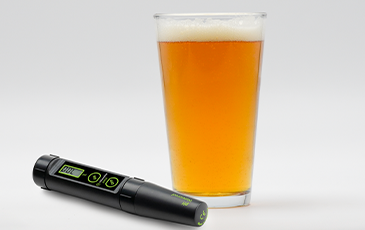 Digital pH meter lying next to a pint glass of golden beer with a foamy head.