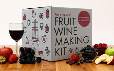 Image of the Fresh Harvest Fruit Wine Making Kit on a wood table with a glass of wine surrounded by different types of fruit. 