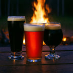 Image of 3 Beers Two dark ales and one red ale on a table in front of a campfire in the woods