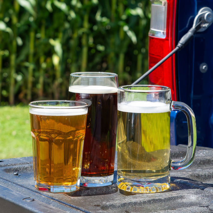 Close-up of three glasses of beer—cream ale, Kentucky common, and golden pre-prohibition lager—on a truck tailgate with a blurred cornfield background.
